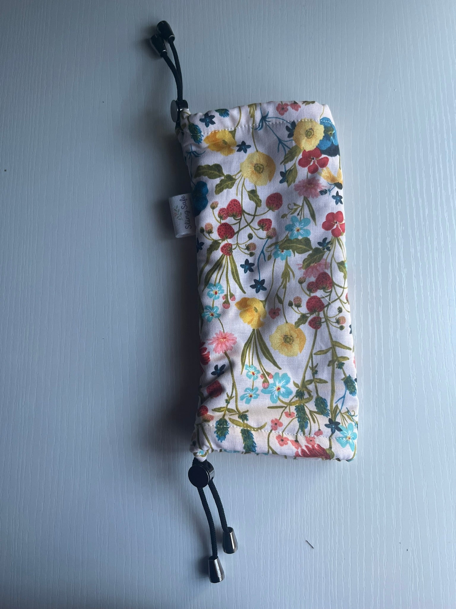 Sling Safe drawstring washing cover for ring slings, shown in the pattern Wildflower. Wildflower is a beige background with multiple flowers and strawberries throughout. 